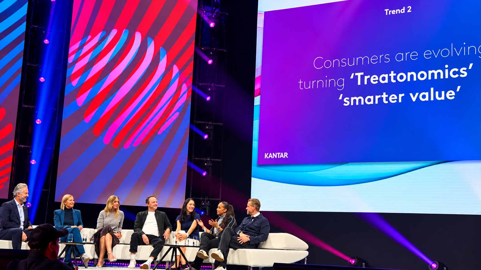 Original-Praesentationsfolie mit dem Text 'Consumers are evolving, turning Treatonomics into smarter value', Trend 2, im Hintergrund der Sprecher auf der Buehne